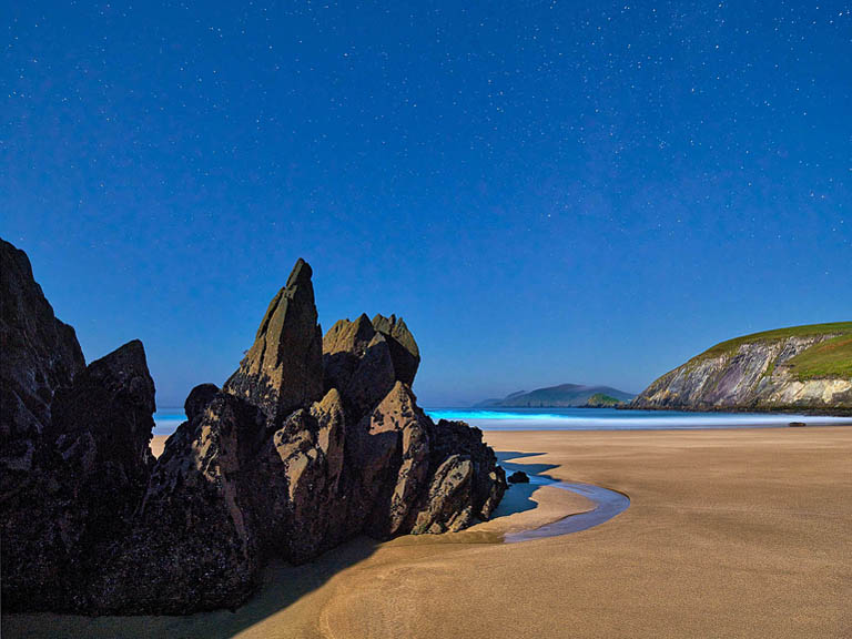 Astrophotography of Coumeenole Beach on the Dingle Peninsula at night, bioluminescent surf and waves rolling in below dark cliffs and a bright sky.