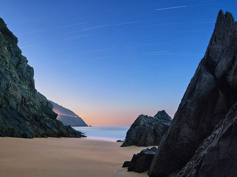 Night-time seascape photograph of Coumeenole Beach on the Dingle Peninsula, surf breaking towards the viewer with cliffs and sky fading into the darkness.