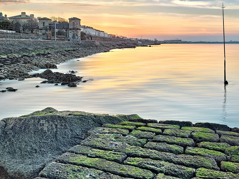 Sunrise beams radiating over the derelict Blackrock Baths and Dart line beside Dublin Bay