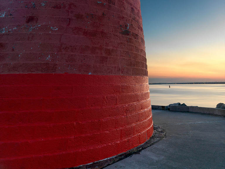 Fine art photograph of Poolbeg Lighthouse on the Great South Wall in Dublin Bay, with red tower, granite wall, sea and sunset sky.