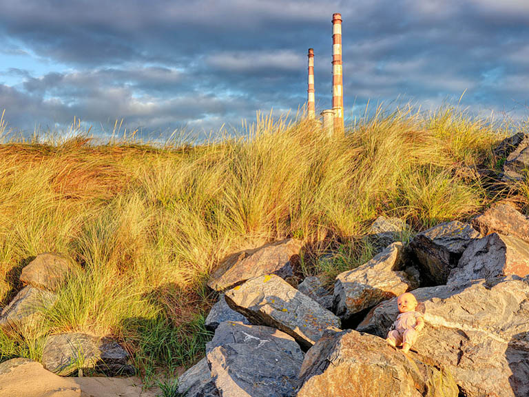 Fine art photograph of a washed up doll on the shore near the Pigeon House in Dublin Bay, with pebbles, seaweed and tide marks.