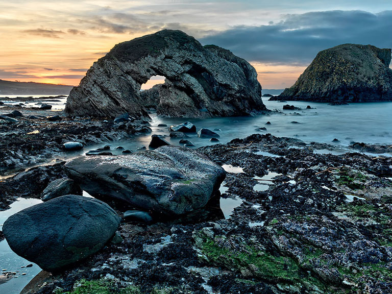 Sunset photograph of Ballintoy Arch on the County Antrim coast with warm sky reflected on the Atlantic and rugged rocks.