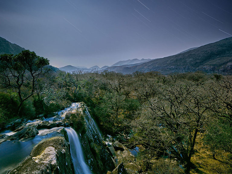 Night photograph of Crinnagh Waterfall in Killarney Forest Park with star trails and the MacGillycuddy’s Reeks in the background