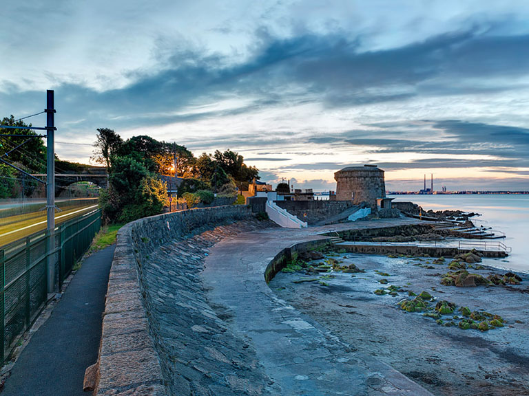 Seascape photograph of Seapoint Martello Tower at dusk over Dublin Bay, with the DART passing in the distance