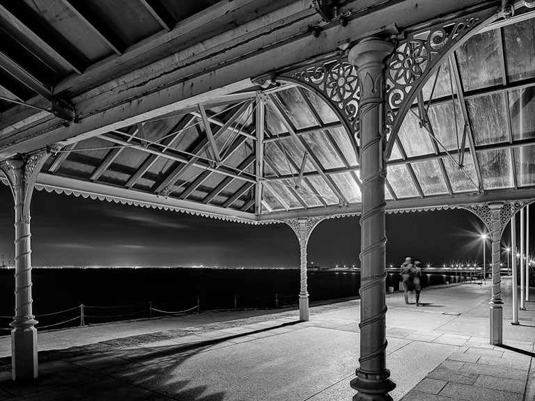Black and white night photograph of the sun shelter and bandstand on Dun Laoghaire East Pier in Dublin, showing wrought iron structure by the sea