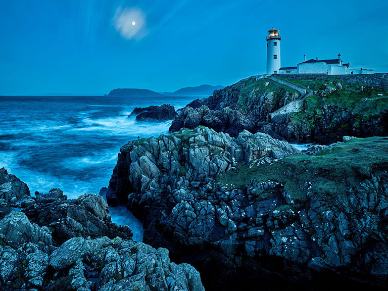Night-time seascape photograph of Fanad Lighthouse above a rocky inlet on the Donegal coast, lit by moonlight