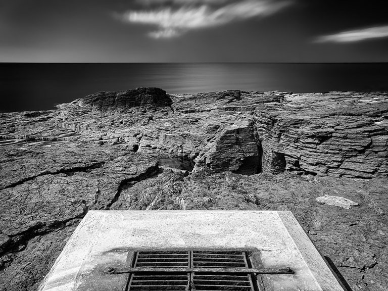 Black and white photograph of a concrete base and steel grille set into rock at Hook Head, County Wexford, with sea beyond