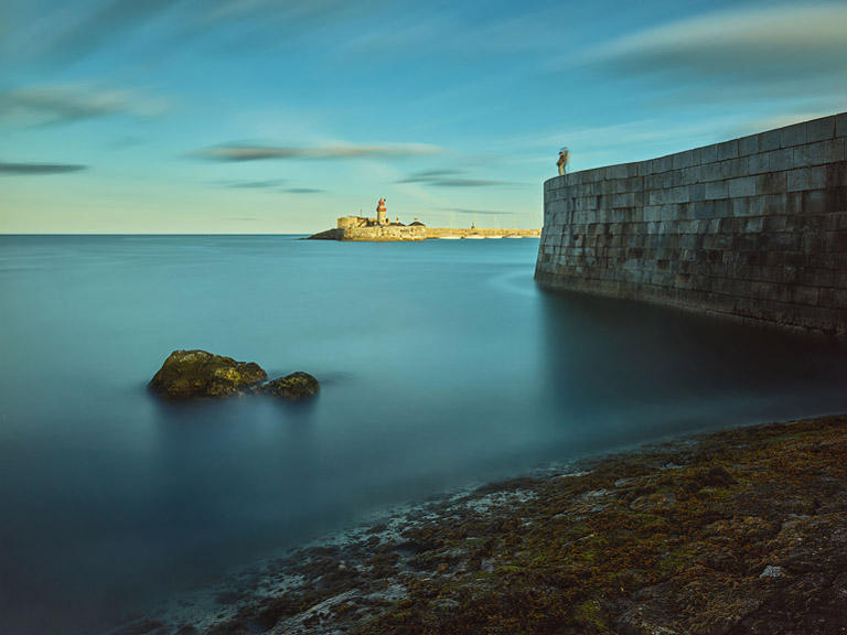 Fine art photograph of Dun Laoghaire West Pier at dusk with calm water and the lights of the East Pier in the distance.