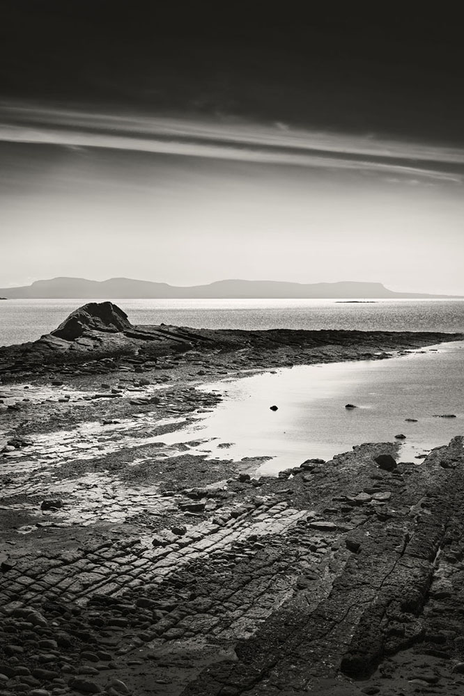 Black and white view of rocky shoreline and calm sea at Rinn Point, County Donegal, with Sligo in the distance
