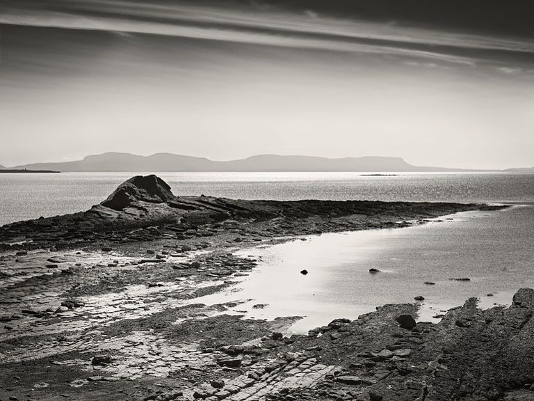 Black and white view of rocky shoreline and calm sea at Rinn Point, County Donegal, with Sligo in the distance