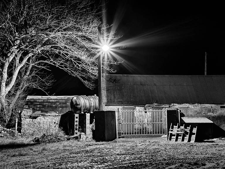 Black and white photograph of the old family farm sheds in Skreen, County Sligo, with trees and a quiet farmyard where the artist played as a child.
