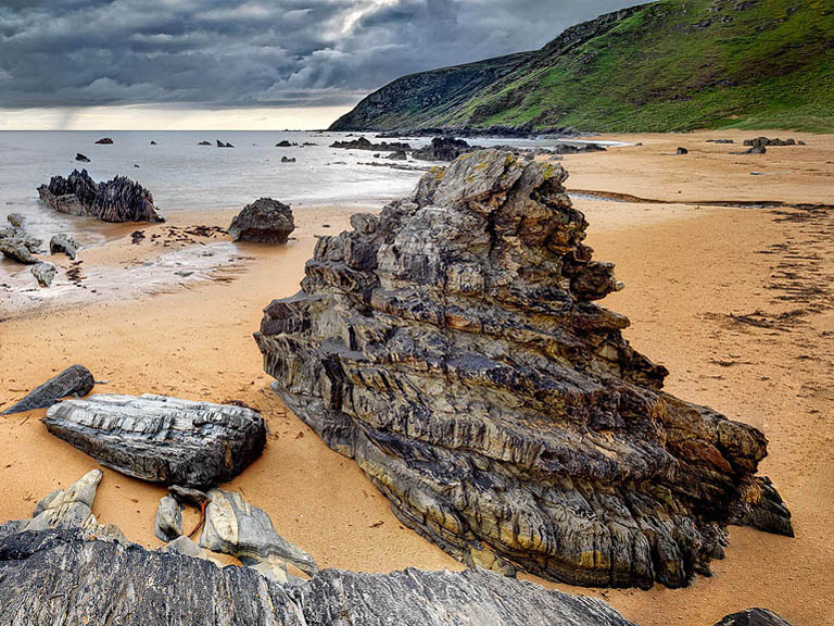 Fine art photograph of dark metallic rock formations on Kinnagoe Beach on the Inishowen Peninsula with the Atlantic surf beyond