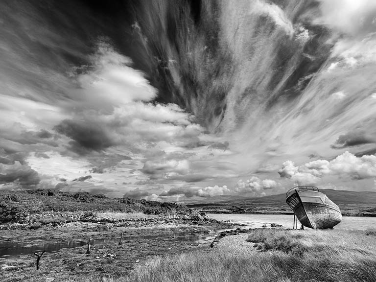Fine art photograph of a ruined wooden boat on the shore of Dunmanus Bay in West Cork, with bay and dramatic clouds behind