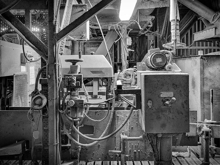 Black and white workplace photograph inside an animal feed mill, with machinery, conveyors and dust-filled light in the interior